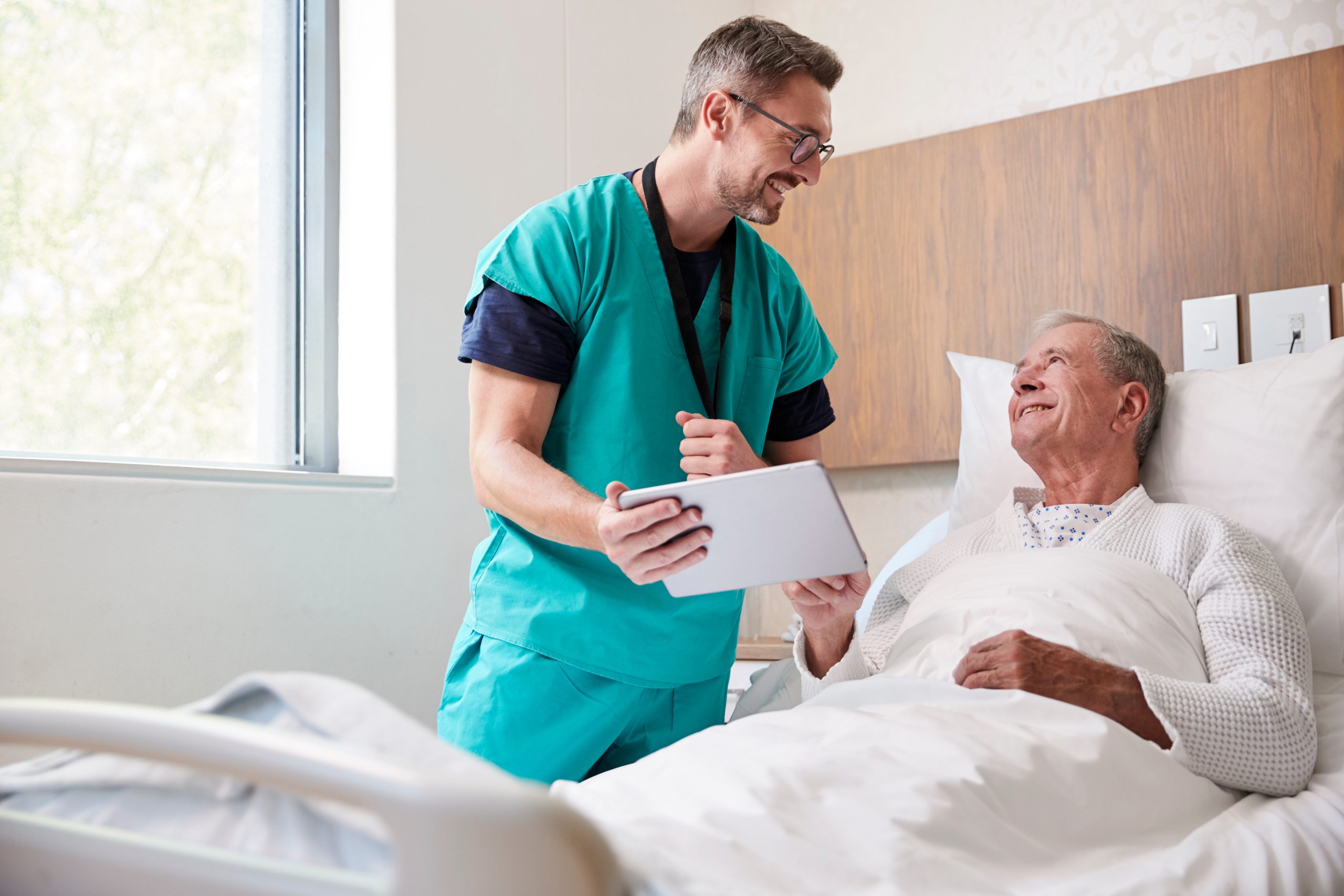 Surgeon With Digital Tablet Visiting Senior Male Patient In Hospital Bed In Geriatric Unit